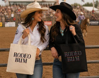 May include: Two canvas tote bags, one cream and one black, each featuring the text "RODEO BUM" in a colorful, multi-striped pattern. The bags have two sturdy handles and a rectangular shape, displayed in a setting with a dirt arena and fencing.