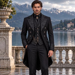 Op de afbeelding: Een zwart rokkostuum met een vest en revers van brokaat met patroon, een zwart overhemd en een gouden horlogeketting. Het ensemble bevat een bijpassende zwarte plastron met een decoratieve speld, gecombineerd met een zwarte pantalon.