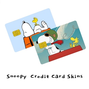 Puede incluir: Dos pegatinas rectangulares para tarjetas de crédito con personajes de Peanuts. Una muestra a Snoopy y Woodstock sobre un fondo azul claro. La otra representa a Snoopy como piloto con bufanda roja y a Woodstock en un tejado rojo. Texto: Snoopy Credit Card Skins.