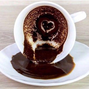 以下が含まれることがあります： 白い陶器製のコーヒーカップとソーサー。カップは傾いており、濃い茶色のコーヒーがソーサーにこぼれています。カップの内側のコーヒーの粉が、白い陶器の表面にくっきりとハートの形を描いています。