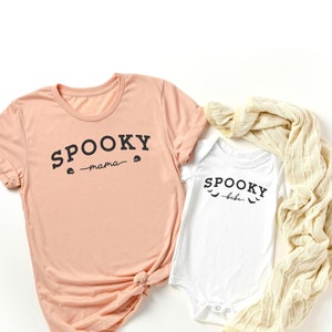 May include: A peach colored t-shirt with the text "SPOOKY -mama- " and a skull graphic. A white onesie with the text "SPOOKY -babe- " and a bat graphic.