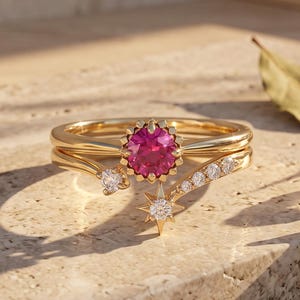Op de afbeelding: Een gouden ring met een centrale rondgeslepen roze edelsteen, geaccentueerd door een stervormige zetting met een kleine heldere steen en een gebogen band versierd met een reeks kleinere heldere stenen, wat een hemels sieraad vormt.
