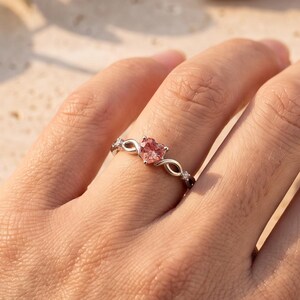 Peut inclure: Une bague argentée ornée d'une pierre précieuse rose en forme de cœur au centre. L'anneau présente un motif en forme d'infini de chaque côté de la pierre, agrémenté de petites pierres claires. La bague est portée sur un doigt.