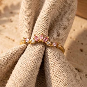 Peut inclure: Une bague dorée présentant un design incurvé en forme de couronne, ornée de pierres roses taille marquise alternant avec de petites pierres rondes transparentes. La bague est placée autour d'un tissu en lin beige plié, révélant son anneau fin.