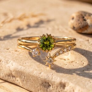 Op de afbeelding: Een gouden ring met een centrale ronde groene peridoot in een stervormige zetting, geaccentueerd door een gebogen band versierd met kleine heldere steentjes en een stervormige bedel met een heldere steen aan de andere kant.