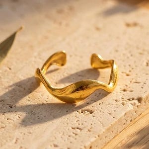 Peut inclure: Une bague dorée présentant un design ouvert avec une bande ondulée et irrégulière. La bague possède une surface polie et réfléchissante qui capte la lumière, créant une forme ondulante distincte qui se courbe autour de l'ouverture.