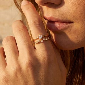Peut inclure: Deux bagues dorées portées sur un doigt. La bague supérieure présente une pierre transparente en forme d'étoile, tandis que la bague inférieure est un anneau incurvé orné d'une série de petites pierres transparentes taillées en rond.