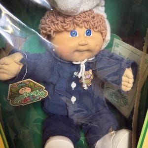 Pode incluir: Uma boneca Cabbage Patch Kids de 1985 com cabelo de lã castanho claro, olhos azuis e covinhas nas bochechas. A boneca usa um gorro de malha branco com um pompom e um pijama de veludo cotelê azul escuro com botões brancos e um emblema de gato.