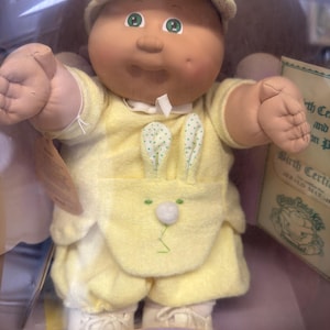 May include: A Cabbage Patch Kids doll from 1985 with a round face, green eyes, and a dimple. The doll wears a yellow cap with green dots, a matching yellow outfit featuring a bunny-themed pocket with a white pom-pom, and white shoes.