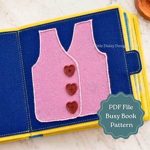 Puede incluir: Una página de libro de actividades de fieltro con dos formas de chaleco rosa claro cosidas sobre un fondo azul oscuro. El chaleco delantero está adornado con tres botones rojo oscuro en forma de corazón. El texto Little Daisy Design Studio y PDF File Busy Book Pattern es visible.