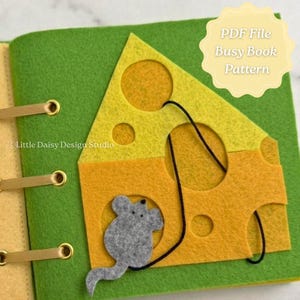 May include: A felt busy book page featuring a yellow cheese wedge with circular cutouts on a green background. A small grey felt mouse is attached to the cheese by a black cord. The text "PDF File Busy Book Pattern" and "Little Daisy Design Studio" are visible.