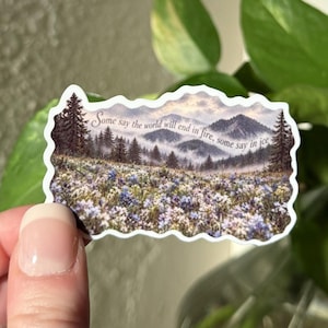 May include: A die-cut sticker featuring a landscape illustration of a field of purple and white wildflowers beneath a mountain range and pine trees. The text "Some say the world will end in fire, some say in ice" is printed across the sky.