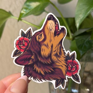 May include: A die-cut sticker featuring a howling wolf head in shades of brown and gold, accented with two vibrant red roses and dark green leaves on either side. The illustration has a bold outline and a detailed, textured fur appearance.