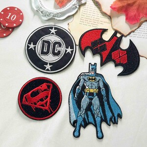 May include: A collection of four embroidered patches featuring a circular black and white DC logo, a black and red bat-shaped emblem, a circular black and red Superman shield, and a full-body depiction of Batman in a blue and grey costume with a cape.