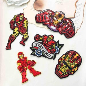 May include: Five embroidered patches featuring Iron Man in various poses and styles. One patch shows the character in a flying pose, another in a standing stance, one in a punching pose, one with the text STAY GOLD, and one helmet design.