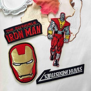 以下が含まれることがあります： マーベルのキャラクターとロゴが刺繍された4つのワッペン。1つは赤と金のアイアンマンのヘルメット、もう1つは赤い文字で「THE INVINCIBLE IRON MAN」、1つはコロッサスのキャラクター、もう1つは「STARK INDUSTRIES」と書かれています。