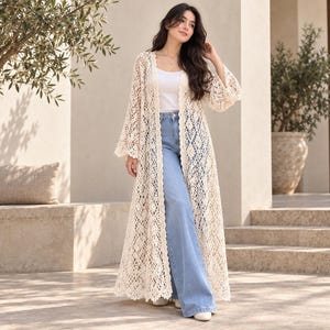 Puede incluir: Un cárdigan largo de ganchillo color crema que llega hasta el suelo, con un intrincado patrón de diamantes calados, mangas largas acampanadas y frente abierto, usado sobre una camiseta blanca sin mangas y jeans azul claro de pierna ancha.