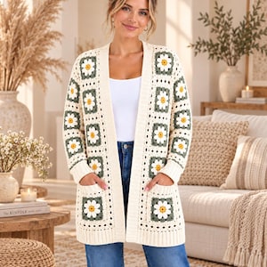 May include: A cream-colored open-front cardigan featuring a repeating granny square pattern with white and yellow daisy motifs on a sage green background. The garment includes long sleeves, ribbed cuffs, a ribbed hem, and two front patch pockets.