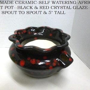 May include: A handmade ceramic self-watering African violet pot with a black and red crystal glaze. The pot features a scalloped rim and a side spout, measuring 9 inches from spout to spout and 5 inches tall as stated in the text.