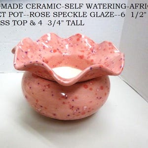 May include: A handmade ceramic self-watering African violet pot with a rose-colored speckled glaze, featuring a ruffled rim. The pot measures 6.5 inches across the top and 4.75 inches tall, as stated in the text on the image.