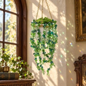 May include: A hanging planter featuring cascading strands of faceted green glass beads in various shades of emerald, lime, and forest green. The beads are suspended from a gold-toned metal chain, creating a shimmering effect with light reflections.