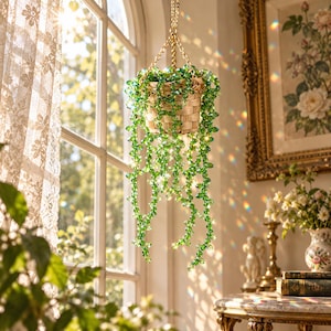 Puede incluir: Una decoración de planta colgante con una cesta tejida de color marrón claro llena de hilos de cuentas de cristal verde facetado. El adorno está suspendido por una cadena de metal dorado que atrapa la luz solar creando reflejos coloridos.