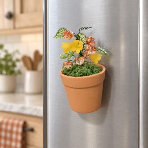 Puede incluir: Una maceta magnética de color terracota que presenta un arreglo decorativo de cuentas translúcidas de color naranja con forma de fruta, flores amarillas en forma de campana y hojas verdes sobre tallos de alambre fino, sobre una base de musgo verde.