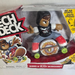 May include: A Tech Deck Shred N' Blast Sk8board toy set in its original packaging. The set includes a stylized figure wearing a black hoodie and blue jeans on a skateboard, along with a ramp. The box features the Tech Deck logo and text.