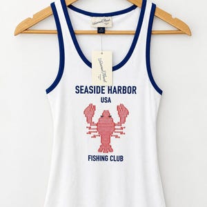 Puede incluir: Una camiseta de tirantes blanca acanalada con ribetes azul oscuro en el cuello y las sisas. El frente presenta un gráfico de langosta roja pixelada con el texto SEASIDE HARBOR USA FISHING CLUB en azul oscuro. Una etiqueta cuelga del cuello.