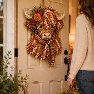 Può includere: Una decorazione da parete a forma di testa di mucca Highland, realizzata con foglie di tessuto stratificate nei toni dell'arancione, marrone e giallo. La mucca presenta due corna color crema, una sciarpa in tessuto scozzese e fiori artificiali.