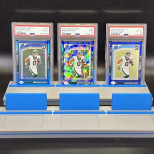 May include: Three 2024 Donruss Optic Braelon Allen trading cards in PSA graded plastic slabs, displayed on a grey and blue tiered plastic stand. The cards feature the athlete in a white uniform, with aqua, ice, and blue glitter parallel designs.