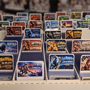 May include: A white cardboard storage box filled with rows of rectangular trading cards, each featuring colorful illustrations of various American cities and sports-themed mascots with text labels like Buffalo, New Orleans, and Los Angeles.
