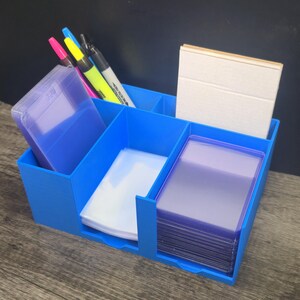 May include: A bright blue multi-compartment desk organizer holding several items. It contains a stack of purple card sleeves, a stack of clear card sleeves, a stack of white cards, and three markers in pink, yellow, and black colors.