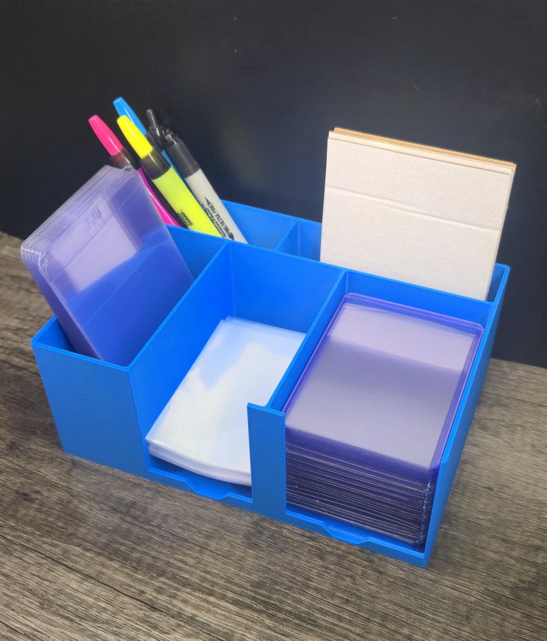 May include: A bright blue multi-compartment desk organizer holding several items. It contains a stack of purple card sleeves, a stack of clear card sleeves, a stack of white cards, and three markers in pink, yellow, and black colors.