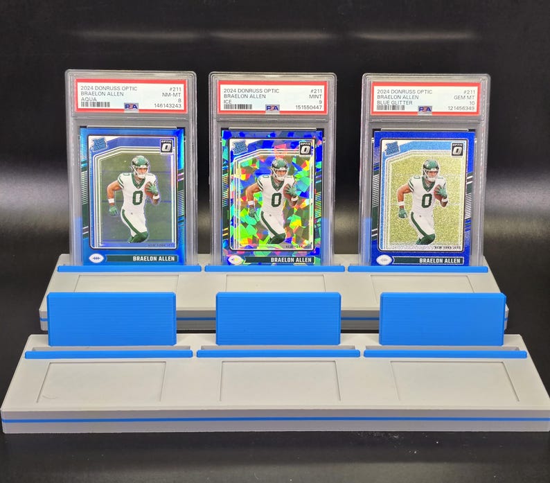 May include: Three 2024 Donruss Optic Braelon Allen trading cards in PSA graded plastic slabs, displayed on a grey and blue tiered plastic stand. The cards feature the athlete in a white uniform, with aqua, ice, and blue glitter parallel designs.