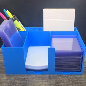 May include: A bright blue plastic desk organizer with multiple compartments. It holds a stack of clear plastic card sleeves, a stack of white paper sheets, a stack of clear rigid card holders, and a set of four colorful markers in the back.