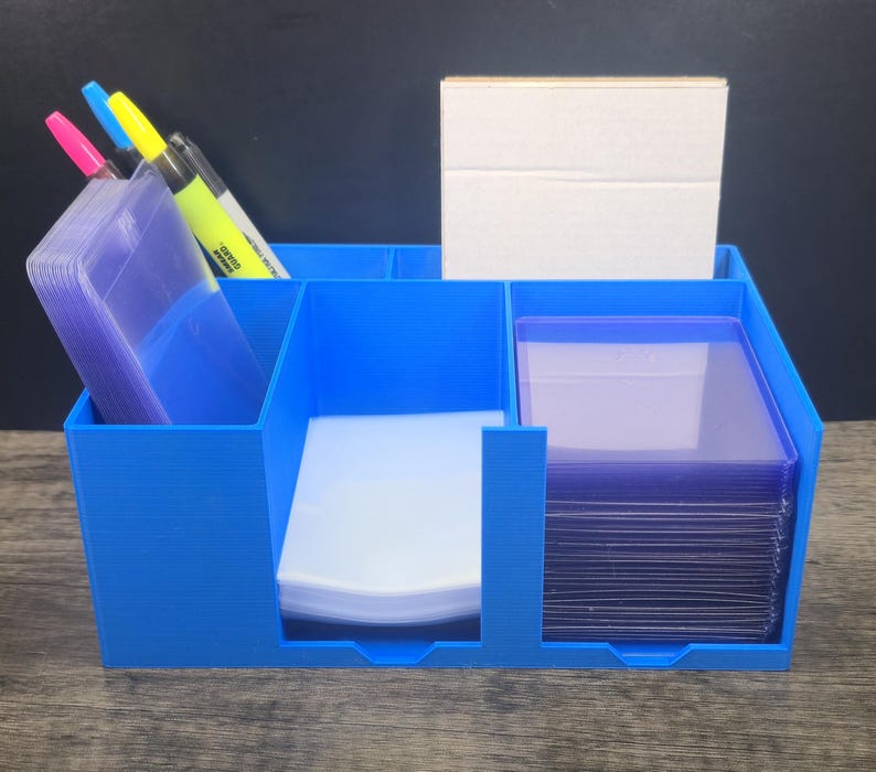 May include: A bright blue plastic desk organizer with multiple compartments. It holds a stack of clear plastic card sleeves, a stack of white paper sheets, a stack of clear rigid card holders, and a set of four colorful markers in the back.