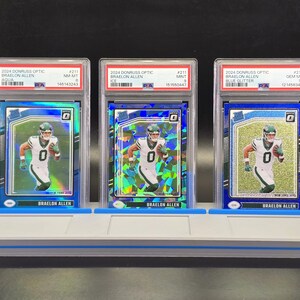 May include: Three 2024 Donruss Optic Braelon Allen football cards in PSA graded plastic slabs, displayed on a grey three-slot stand. Each card features the player in a white jersey with the number zero, showcasing Aqua, Ice, and Blue Glitter parallels.