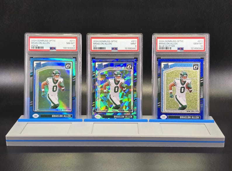 May include: Three 2024 Donruss Optic Braelon Allen football cards in PSA graded plastic slabs, displayed on a grey three-slot stand. Each card features the player in a white jersey with the number zero, showcasing Aqua, Ice, and Blue Glitter parallels.