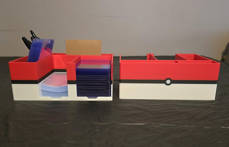 Puede incluir: Un organizador de almacenamiento de pl&aacute;stico rojo, blanco y negro con m&uacute;ltiples compartimentos. El dise&ntilde;o presenta una franja negra horizontal con un c&iacute;rculo blanco central, imitando una bola. Una secci&oacute;n contiene fundas de cartas azules.