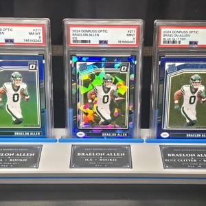 May include: Three 2024 Donruss Optic Braelon Allen football cards in PSA graded slabs, displayed on a light grey stand. The cards feature the player in a white New York Jets uniform, with Aqua, Ice, and Blue Glitter parallel designs and labels.