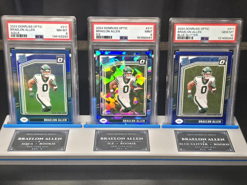 May include: Three 2024 Donruss Optic Braelon Allen football cards in PSA graded slabs, displayed on a light grey stand. The cards feature the player in a white New York Jets uniform, with Aqua, Ice, and Blue Glitter parallel designs and labels.