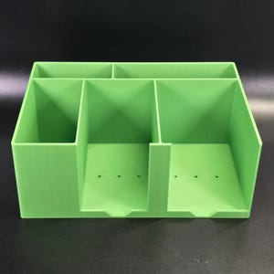 May include: A bright lime green desk organizer featuring a multi-compartment design. The unit includes two wide front slots with small circular drainage holes at the base and two narrower rear storage sections, all constructed with smooth, solid walls.