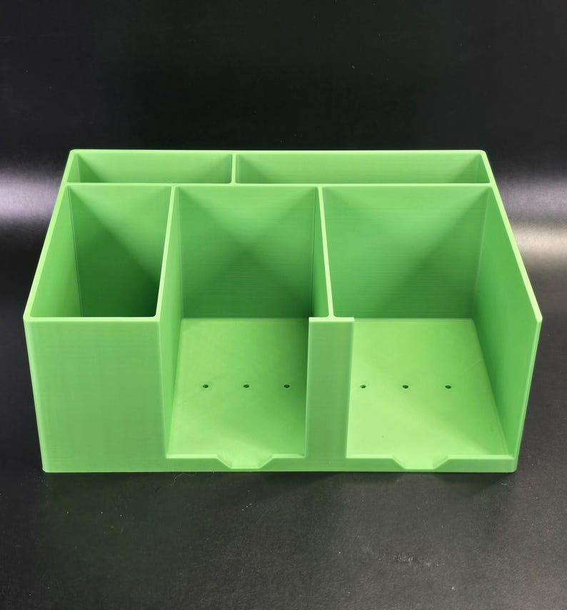 May include: A bright lime green desk organizer featuring a multi-compartment design. The unit includes two wide front slots with small circular drainage holes at the base and two narrower rear storage sections, all constructed with smooth, solid walls.