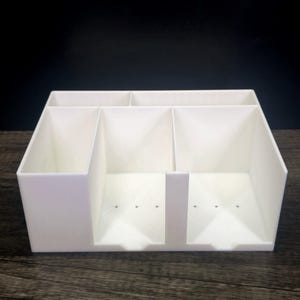 May include: A white plastic desk organizer featuring a rectangular shape with multiple compartments. The unit includes two narrow slots at the back and two larger, deeper front sections with small drainage holes at the base of each compartment.