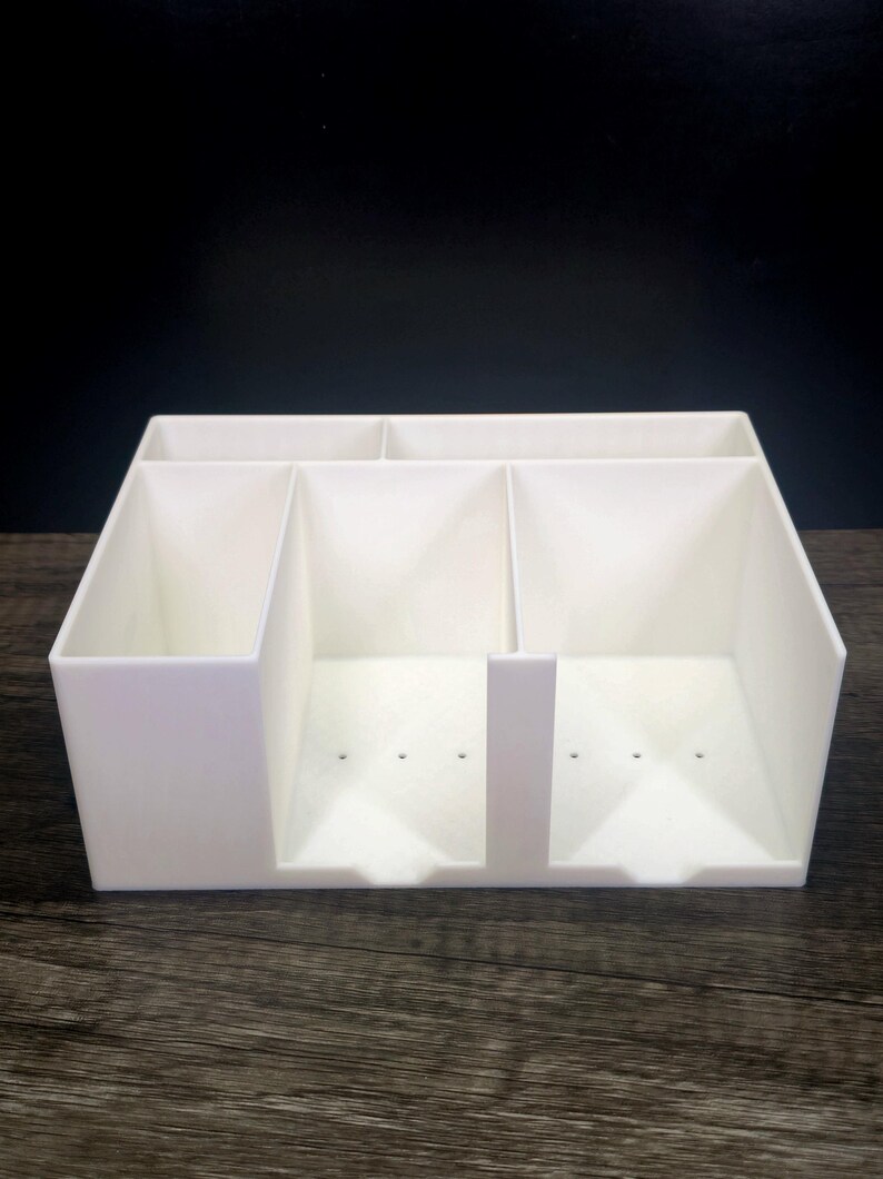May include: A white plastic desk organizer featuring a rectangular shape with multiple compartments. The unit includes two narrow slots at the back and two larger, deeper front sections with small drainage holes at the base of each compartment.