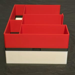 Puede incluir: Un organizador de escritorio rectangular que presenta una secci&oacute;n superior roja con m&uacute;ltiples compartimentos y una secci&oacute;n inferior blanca, separadas por una fina banda horizontal negra. La estructura est&aacute; dividida en varios espacios abiertos.