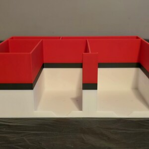 Puede incluir: Un organizador de escritorio rectangular con una secci&oacute;n superior roja y una secci&oacute;n inferior blanca, separadas por una franja negra horizontal. El organizador cuenta con m&uacute;ltiples compartimentos interiores de varios tama&ntilde;os.