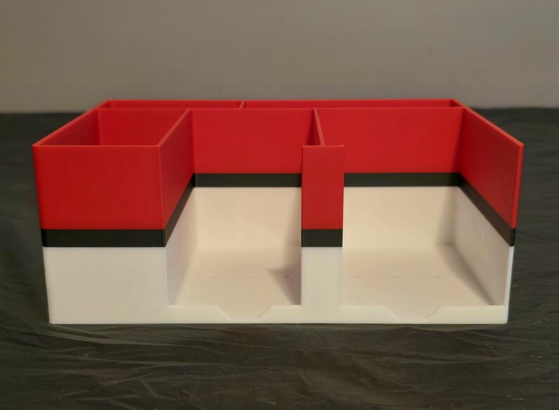 Puede incluir: Un organizador de escritorio rectangular con una secci&oacute;n superior roja y una secci&oacute;n inferior blanca, separadas por una franja negra horizontal. El organizador cuenta con m&uacute;ltiples compartimentos interiores de varios tama&ntilde;os.