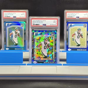 May include: Three graded 2024 Donruss Optic Braelon Allen football cards in PSA plastic cases, displayed on a light grey tiered stand with blue accents. The cards feature the athlete in a white and green uniform, with aqua, ice, and blue glitter finishes.